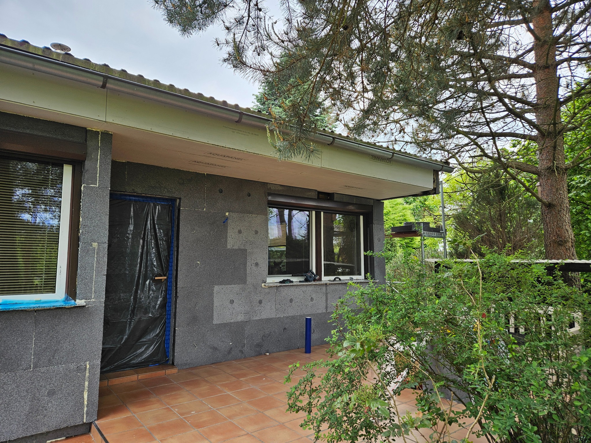 Budynek w trakcie prac związanych z izolacją termiczną, widoczne szare płyty izolacyjne na elewacji, okno zabezpieczone taśmą, a drzwi folią ochronną, na pierwszym planie taras z terakoty i zieleń.