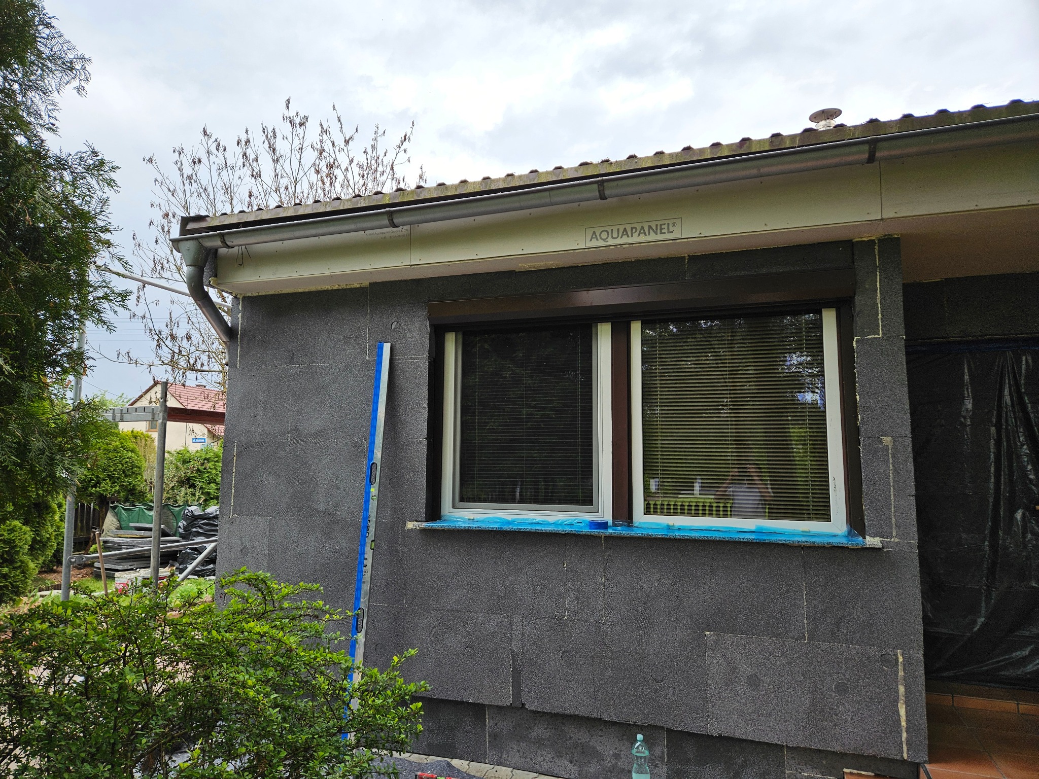 Ocieplanie elewacji budynku płytami AQUAPANEL, widoczna poziomica, okno zabezpieczone folią, prace w toku.