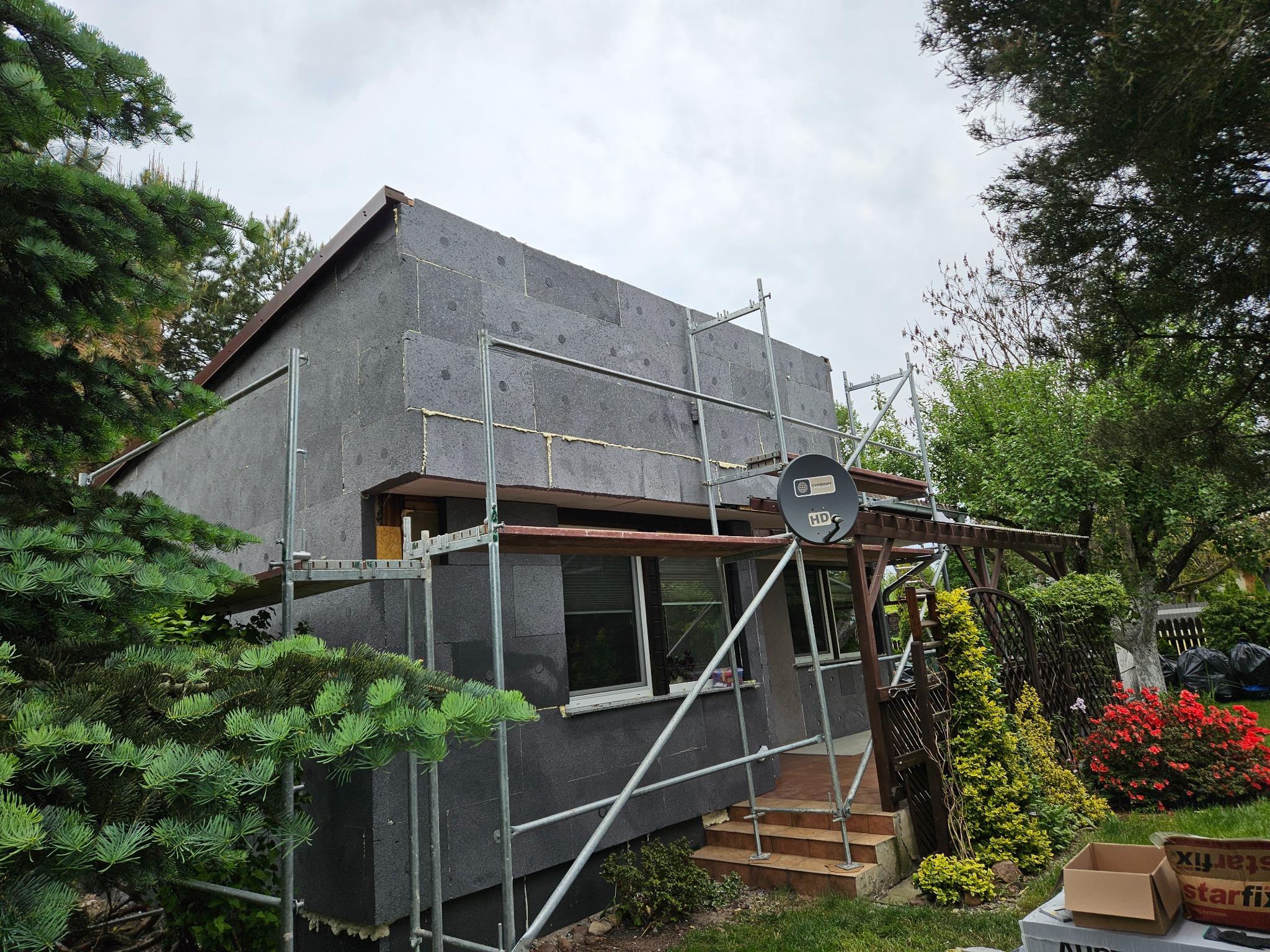 Dom jednorodzinny w trakcie prac, na elewacji szare płyty izolacyjne, widoczne rusztowania, fragment ogrodu z zielenią i kwiatami, antena satelitarna na ścianie.