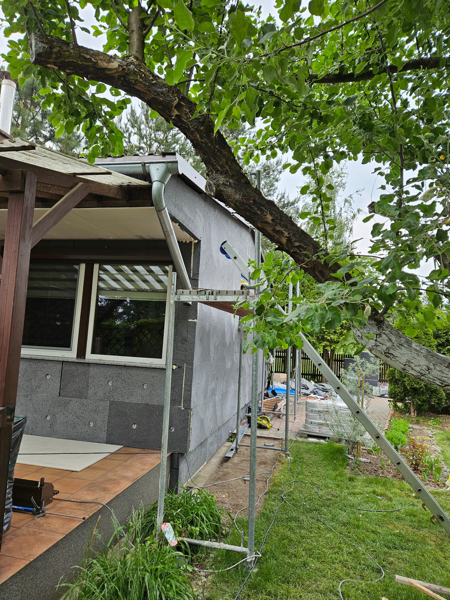 Dom w trakcie ocieplania elewacji, widoczne szare płyty styropianowe, rusztowanie, drabina oparta o ścianę, oraz gałąź drzewa zwisająca nad budynkiem.