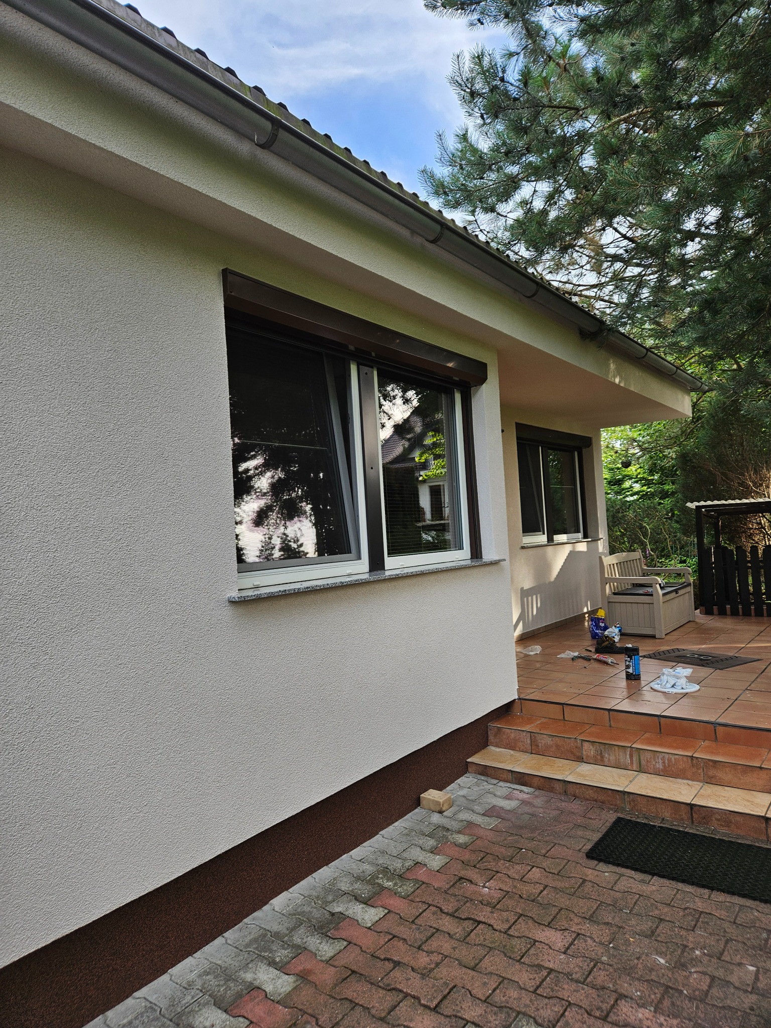 Jasna elewacja domu jednorodzinnego z brązowymi roletami zewnętrznymi i wykończeniem, widoczne narzędzia i materiały budowlane na tarasie, otoczenie zieleni.