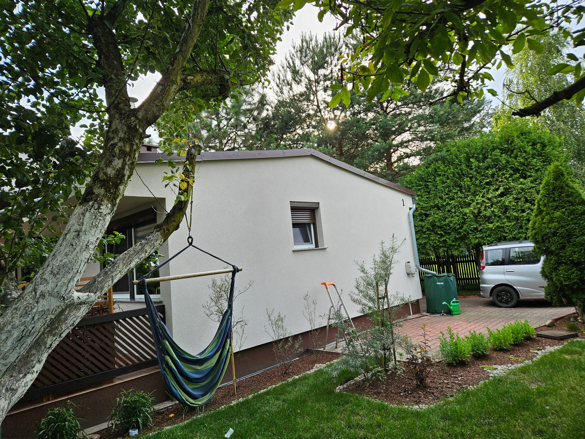 Biały dom jednorodzinny z ocieploną elewacją, widoczny z ogrodu, z hamakiem zawieszonym na drzewie i drabiną opartą o ścianę, sugerującą prace wykończeniowe na zewnątrz.