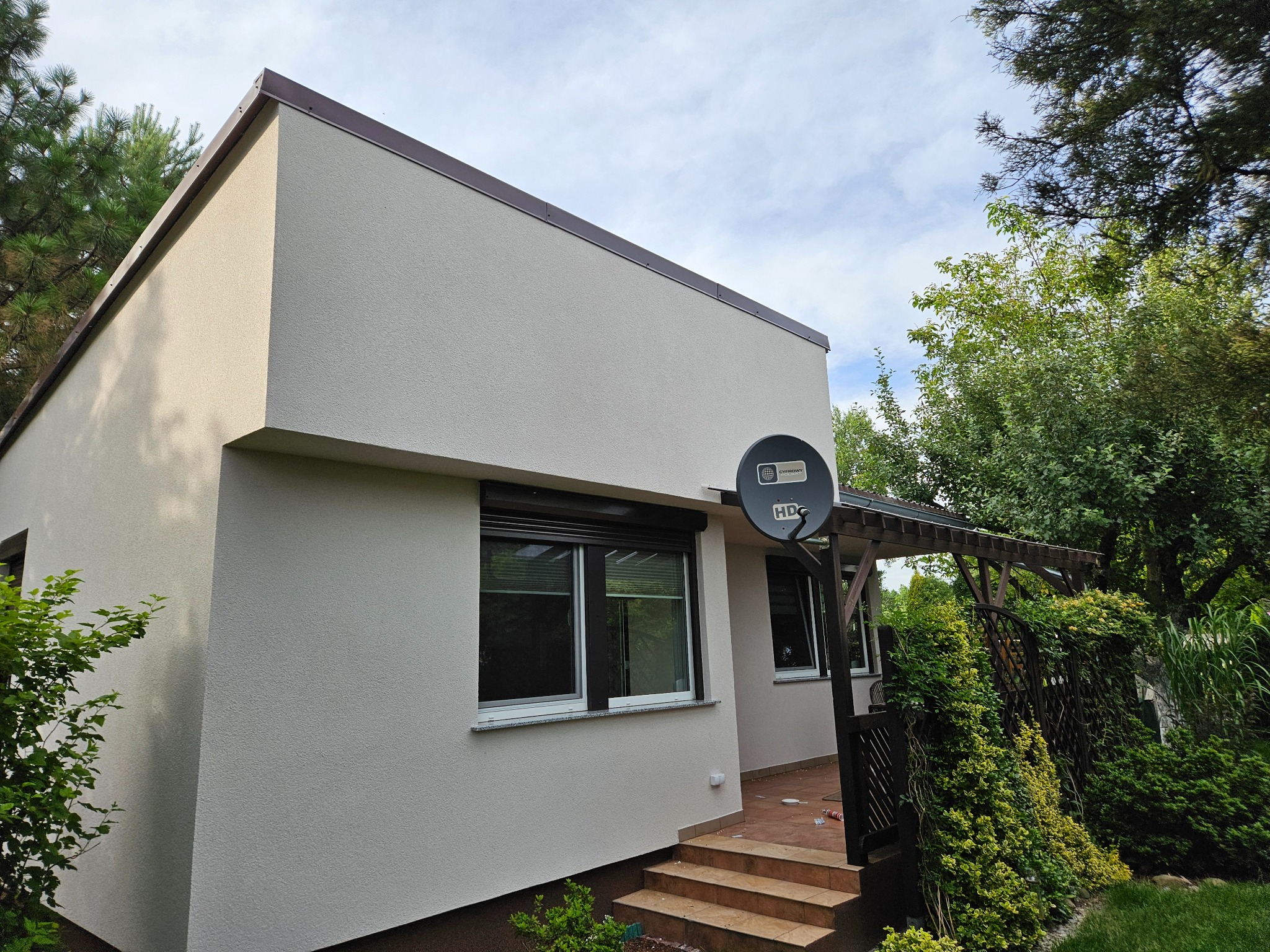 Ocieplona elewacja domu jednorodzinnego z brązowym orynnowaniem i anteną satelitarną, widok z boku na wejście z drewnianymi schodami i pergolą porośniętą zielenią.