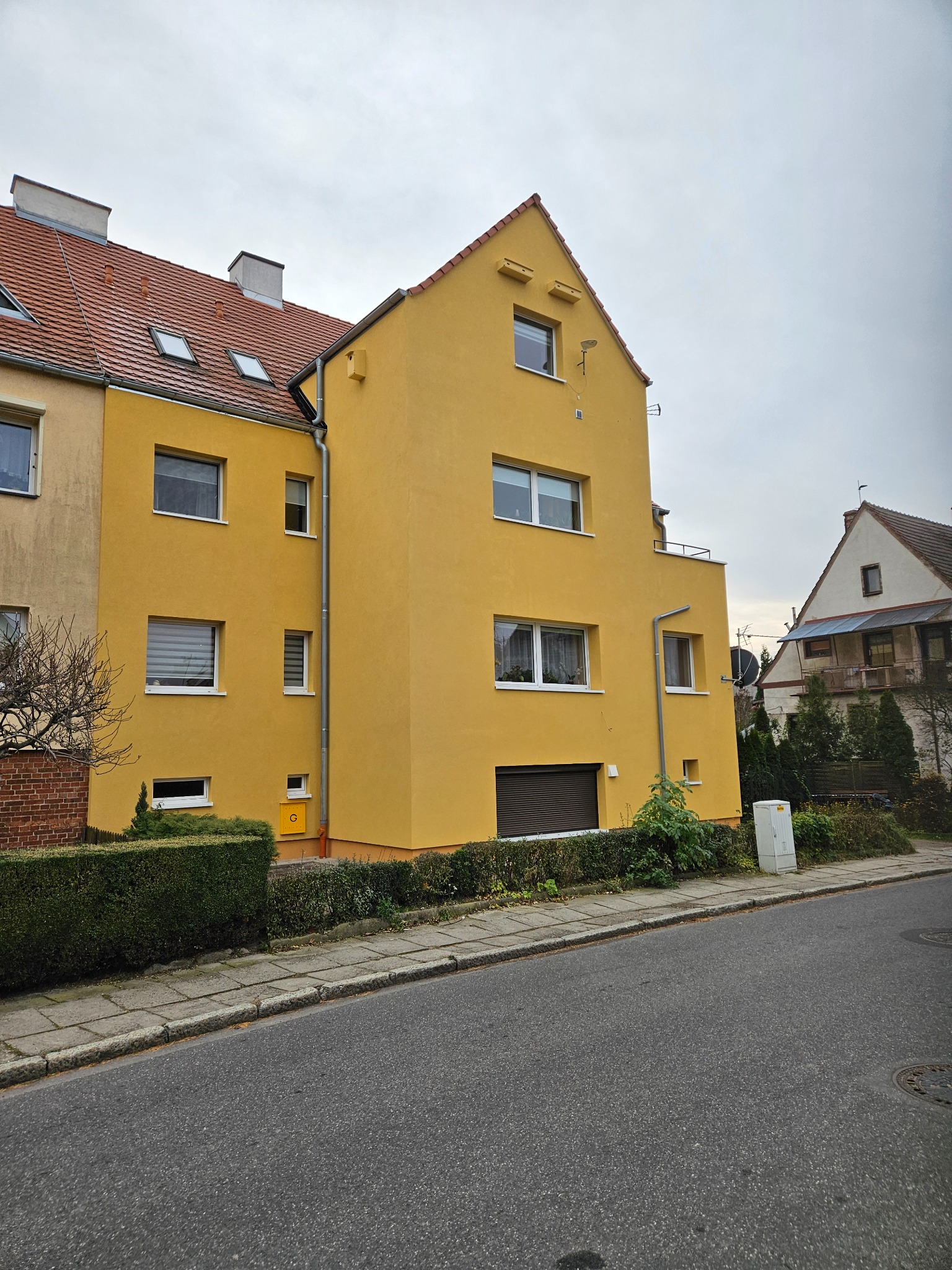 Żółta elewacja odnowionego budynku mieszkalnego z brązową roletą, widoczna z poziomu ulicy w pochmurny dzień. Krzewy wzdłuż chodnika.