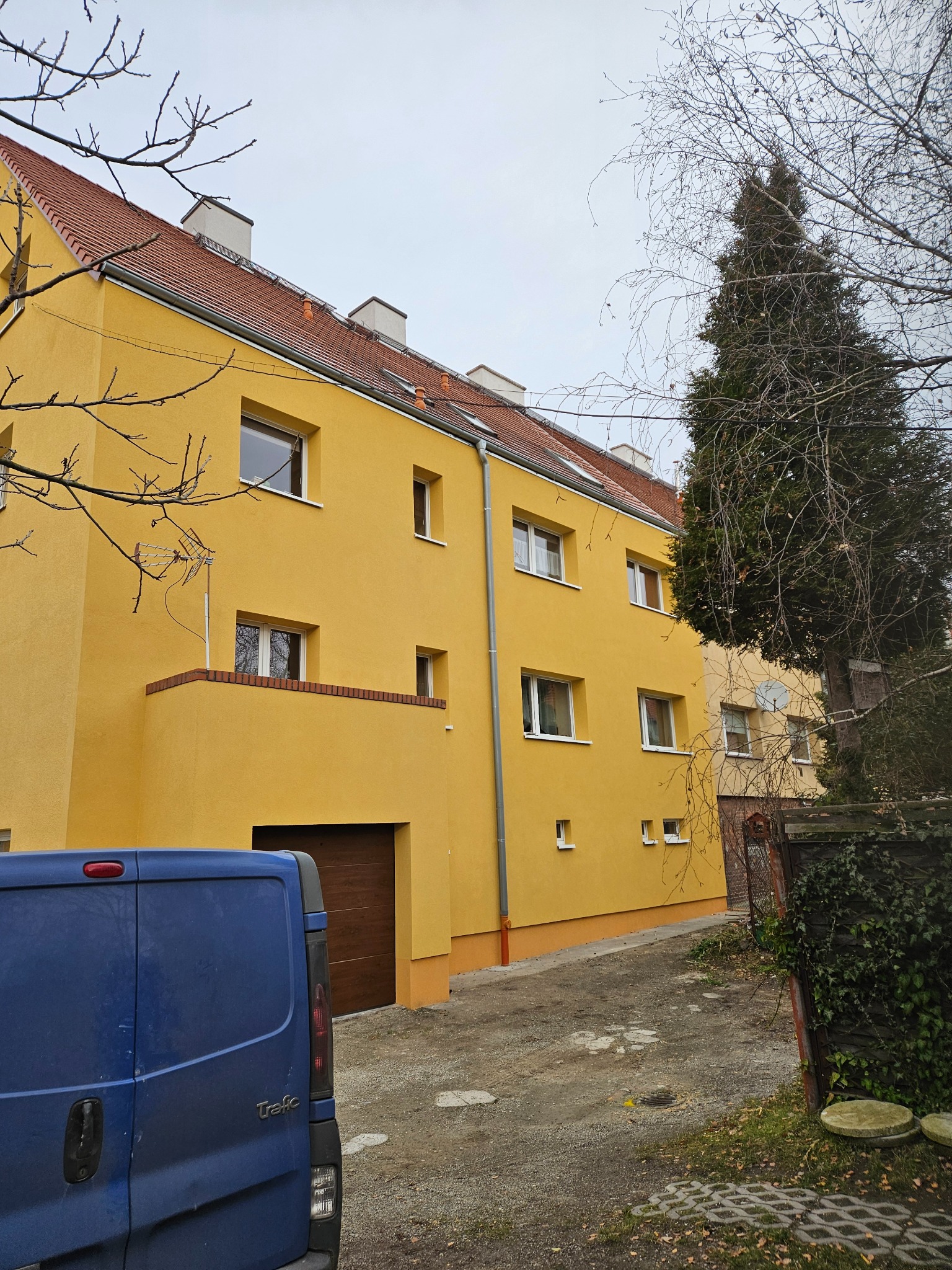 Żółty budynek wielorodzinny z czerwonym dachem, widoczny z perspektywy ulicy. Na pierwszym planie fragment niebieskiego samochodu dostawczego.