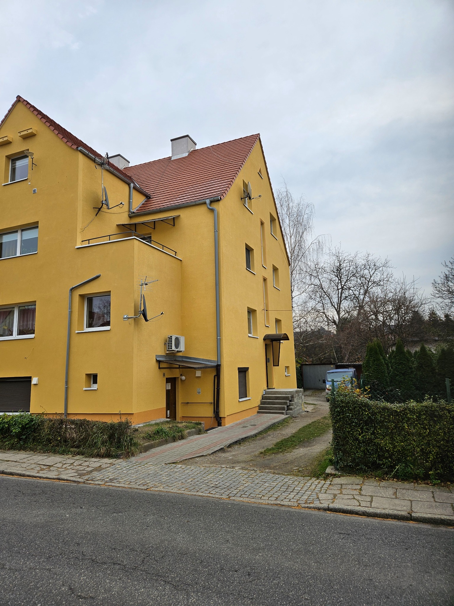 Żółty budynek wielorodzinny z czerwoną dachówką, widoczny z poziomu ulicy, z antenami satelitarnymi na elewacji i chodnikiem prowadzącym do wejścia.