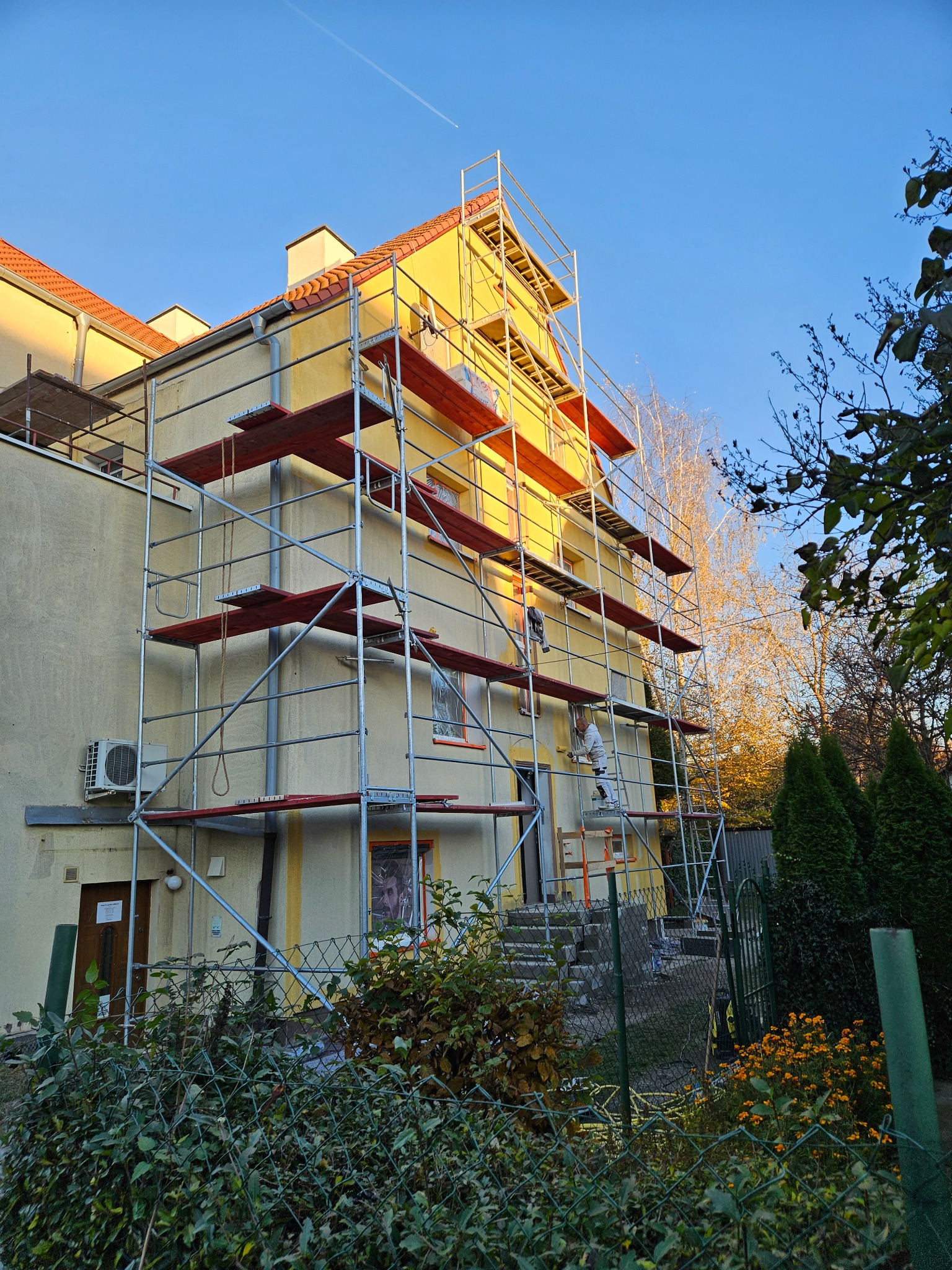 Żółty budynek w trakcie prac elewacyjnych, otoczony rusztowaniem. Pracownik w białym kombinezonie maluje ścianę. Widoczne drzewa i krzewy w otoczeniu.