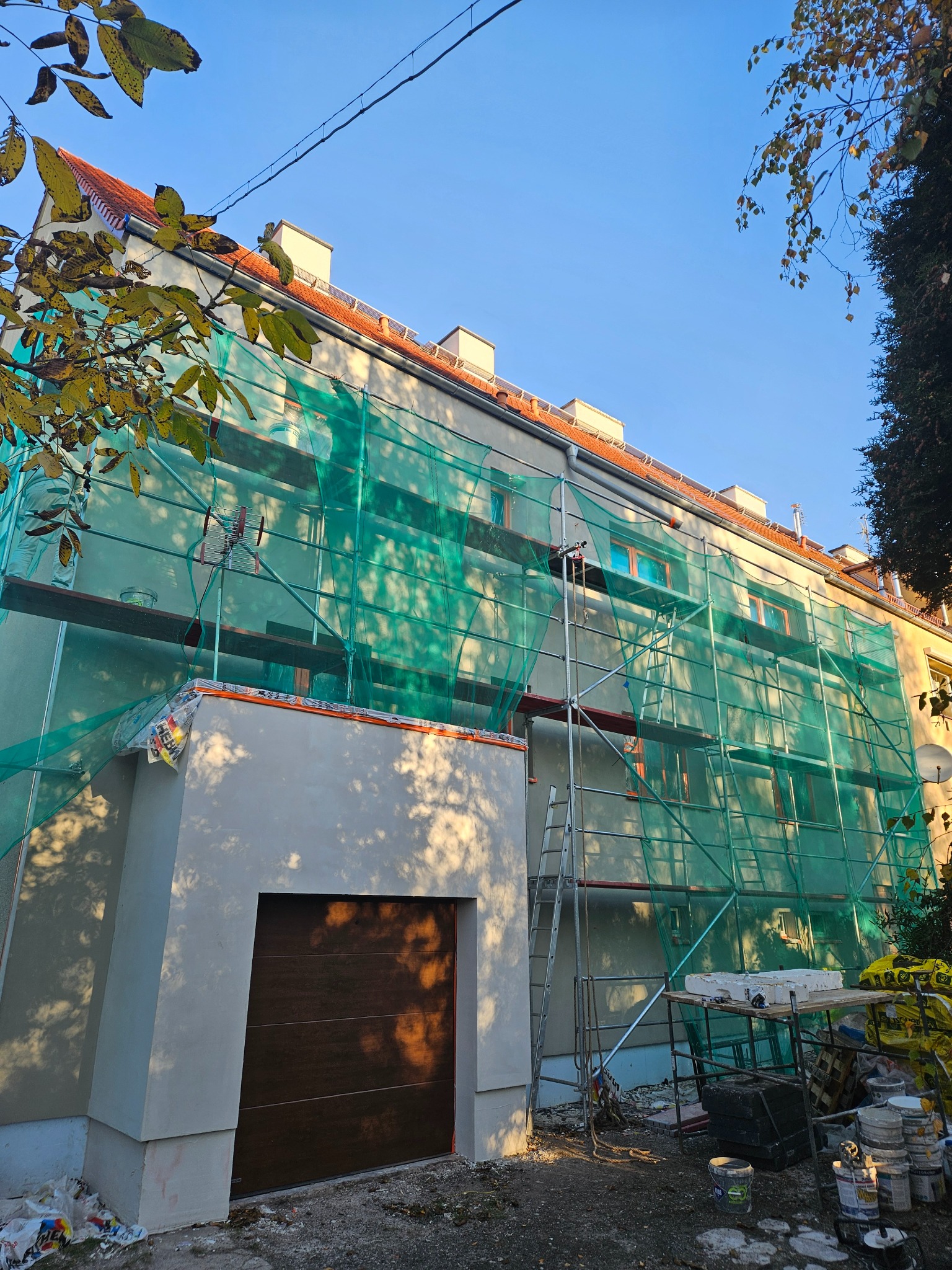 Budynek w trakcie prac elewacyjnych, osłonięty siatką ochronną, widoczne rusztowania i świeżo otynkowany garaż z brązową bramą. Słoneczny dzień.