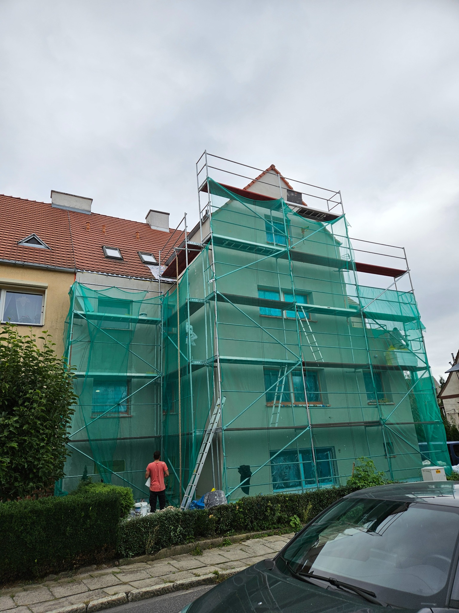 Budynek w trakcie prac elewacyjnych, osłonięty siatką ochronną na rusztowaniu. Widoczny pracownik w pomarańczowej koszulce i wiadro z farbą. Ujęcie z dołu, pochmurne niebo.