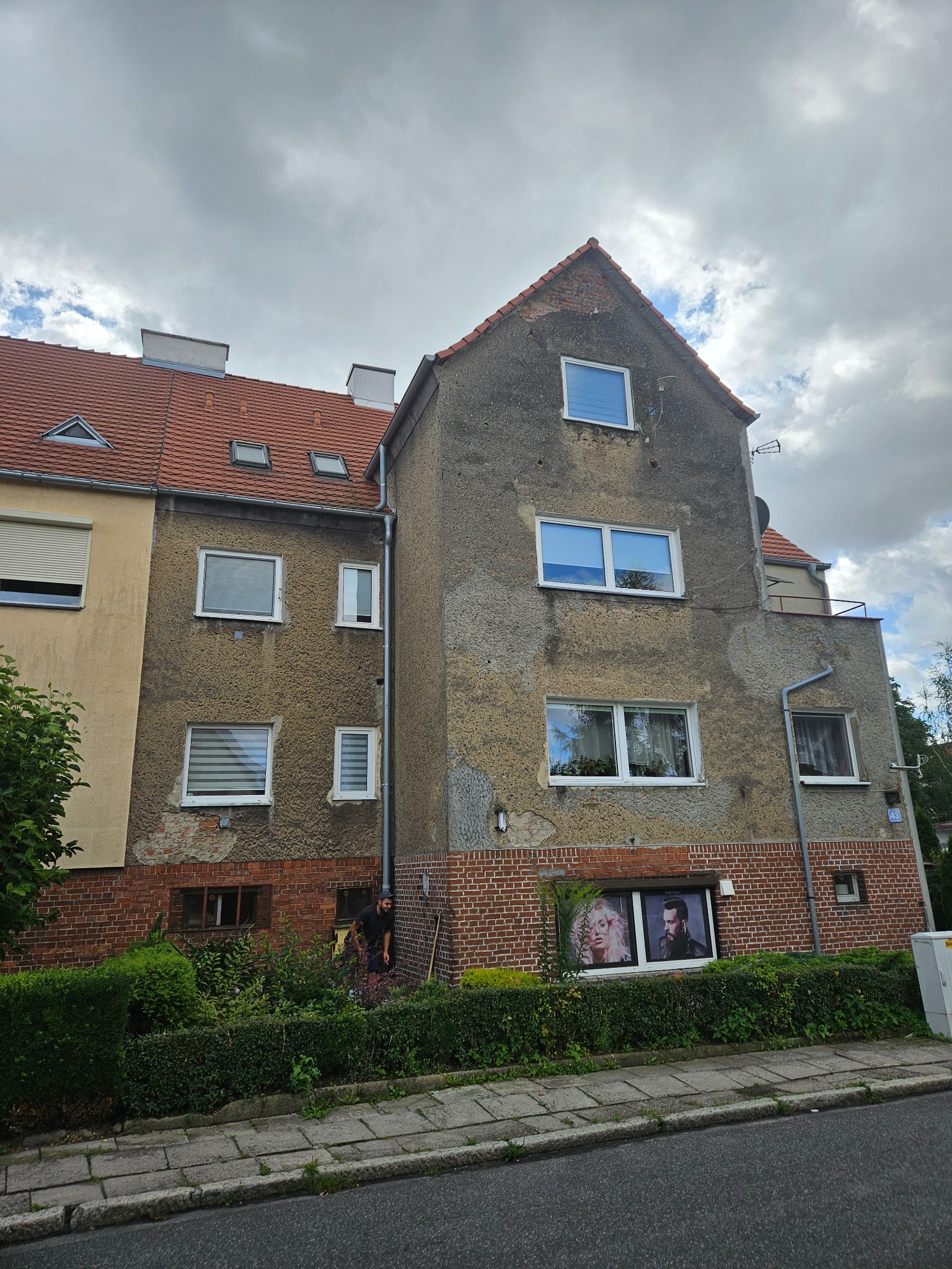 Dwupiętrowy budynek z widocznymi śladami uszkodzeń elewacji, z czerwonym dachem i oknami. Na parterze okna z reklamami. Osoba przy budynku. Pochmurne niebo.