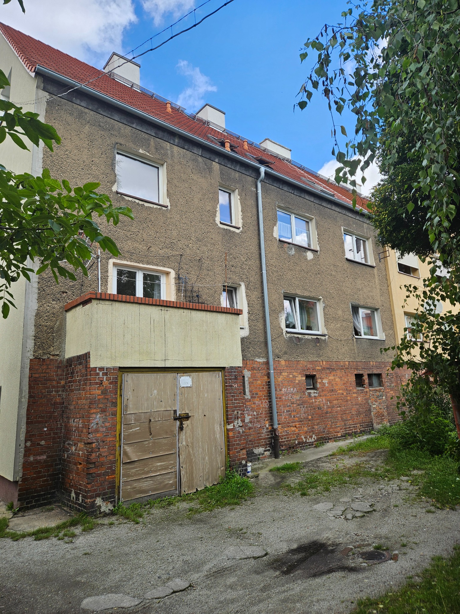 Szary budynek wielorodzinny z czerwonym dachem, widoczny z dołu. Na parterze ceglana podmurówka i drewniane drzwi garażowe. Fragmenty zieleni po bokach.