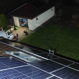 Krzysztof Szymecki - Widok z góry na panele fotowoltaiczne na dachu domu jednorodzinnego nocą; w tle widoczny garaż i częściowo samochód dostawczy z wyposażeniem.