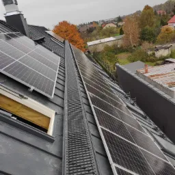 Panele słoneczne zamontowane na ciemnym dachu skośnym, widoczna drabinka dachowa i komin, w tle jesienny krajobraz.