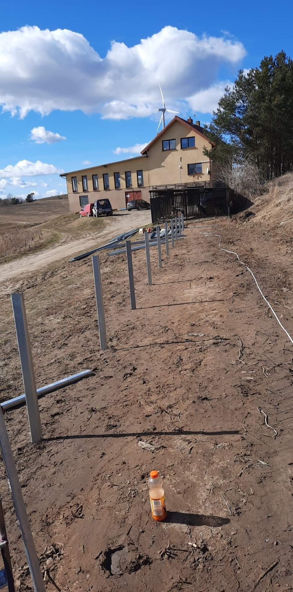 Przygotowanie terenu pod instalację paneli fotowoltaicznych: wbite metalowe słupki w ziemię, w tle budynek z wiatrakiem, butelka z napojem na pierwszym planie.