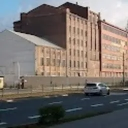 Duży, ceglany budynek przemysłowy widziany z ulicy. W tle błękitne niebo. Na pierwszym planie fragment drogi i zaparkowany samochód osobowy.