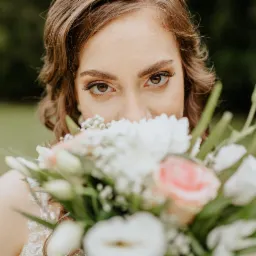 Portret panny młodej z bliska, z bukietem kwiat&oacute;w zasłaniającym usta, skupiający się na wyrazistych oczach z delikatnym makijażem i kręconych włosach.