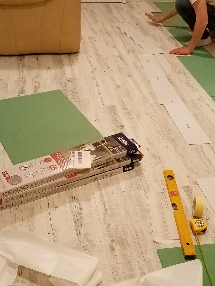 Prace przygotowawcze do ułożenia paneli podłogowych Senso Rustic w mieszkaniu w Warszawie. Widoczne rozłożone panele, paczki z panelami, poziomica, miarka i taśma klejąca.