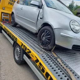 Srebrny, uszkodzony Volkswagen Polo na żółtej lawecie pomocy drogowej z widocznymi numerami telefonu. Uszkodzenia karoserii widoczne na przednim błotniku i drzwiach.