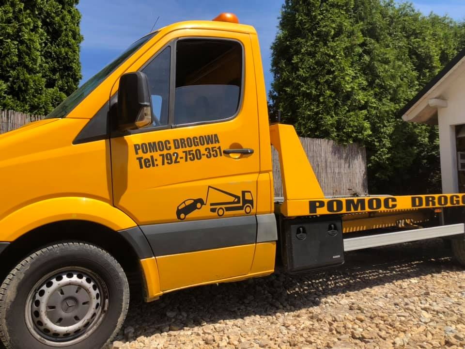 Żółta laweta z czarnym napisem 'POMOC DROGOWA' i numerem telefonu, zaparkowana na żwirowym podłożu, z zielonymi krzewami w tle.