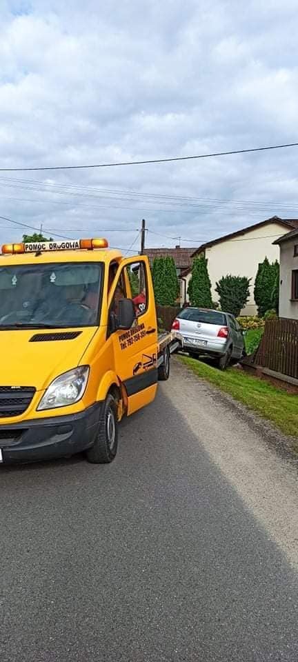 Żółta laweta z włączonymi światłami ostrzegawczymi przewozi srebrny samochód osobowy na tle domów jednorodzinnych i pochmurnego nieba.