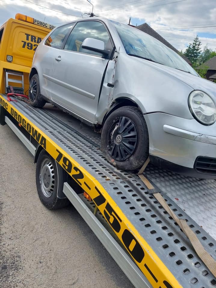 Srebrny, uszkodzony Volkswagen Polo na żółtej lawecie pomocy drogowej z widocznymi numerami telefonu. Uszkodzenia karoserii widoczne na przednim błotniku i drzwiach.