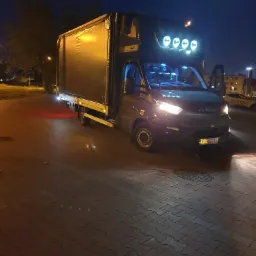 Ciemny bus dostawczy Iveco z plandeką, oświetlony reflektorami dachowymi, stoi na brukowanym placu nocą; widoczne otwarte drzwi kierowcy i tablica rejestracyjna.