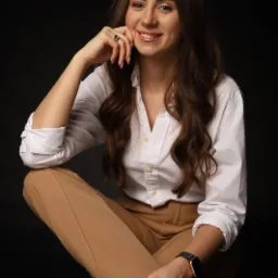 Portret uśmiechniętej kobiety w białej koszuli i beżowych spodniach, siedzącej na czarnym tle, z pierścionkiem z ciemnym oczkiem i zegarkiem na nadgarstku.