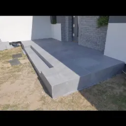 Wykończony taras z szarych płytek ceramicznych przed wejściem do domu, widoczne schody z płytek i obrzeża, trawnik w tle.