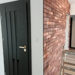 Ciemne drzwi z złotą klamką sąsiadują z fragmentem ściany wyłożonej czerwoną cegłą, widoczna drabina malarska.