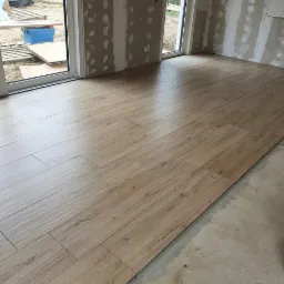 Wnętrze w trakcie układania podłogi z paneli imitujących drewno, widoczne szpachlowane ściany i okno z widokiem na ogr&oacute;d. Część podłogi jeszcze nieułożona.