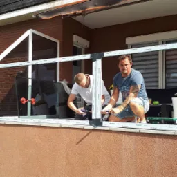 Dwóch mężczyzn montuje szklaną taflę w ramie na balkonie, używając przyssawek do szkła, widoczny profil ramy okiennej z logo producenta.