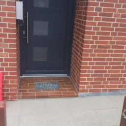 Ciemnoszara, nowoczesna stolarka drzwiowa z trzema szybkami, zamontowana w elewacji z czerwonej cegły z numerem domu.