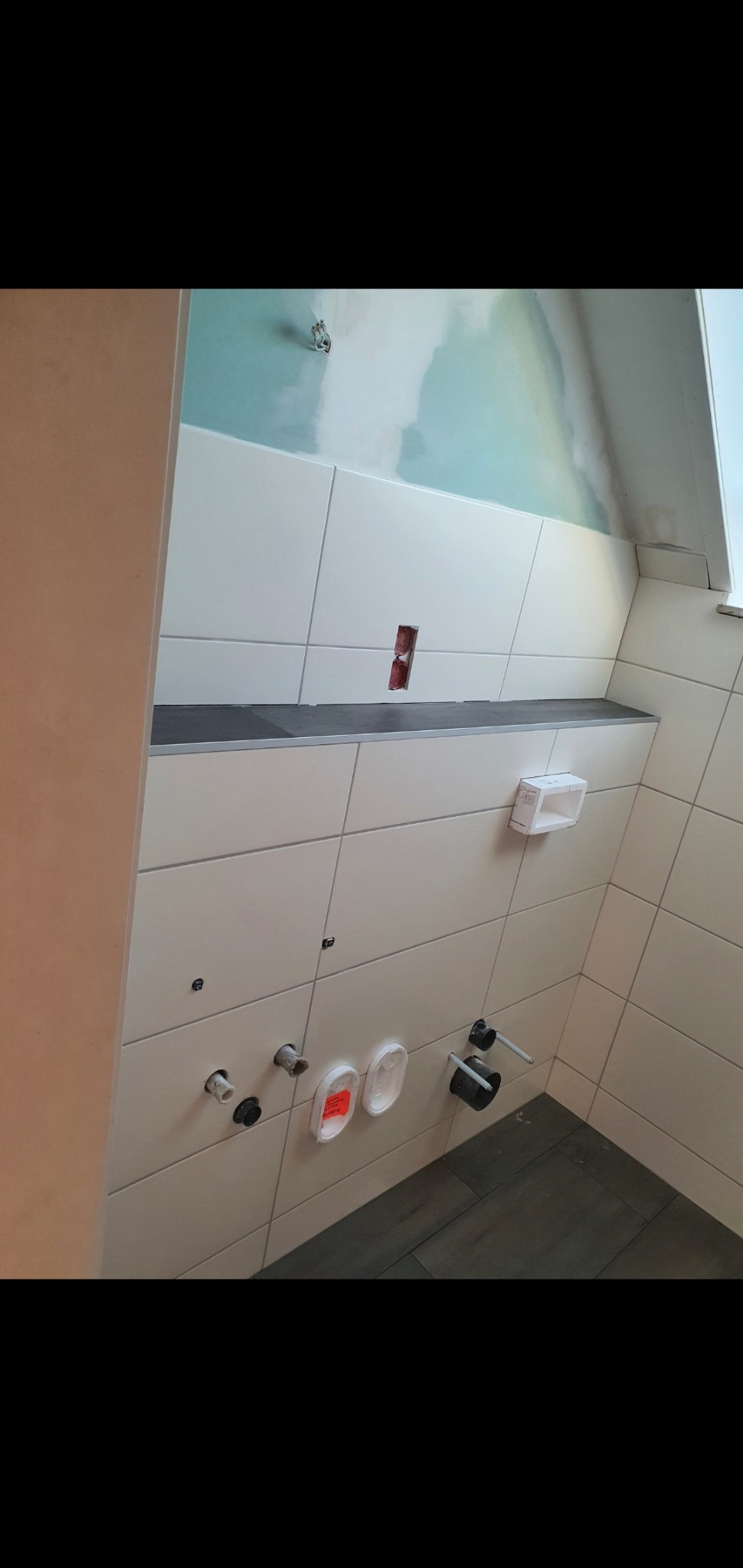 Świeżo wykafelkowana ściana w łazience z przygotowanymi przyłączami wodno-kanalizacyjnymi, widoczny fragment sufitu ze śladami gładzi i niedokończone fugi.