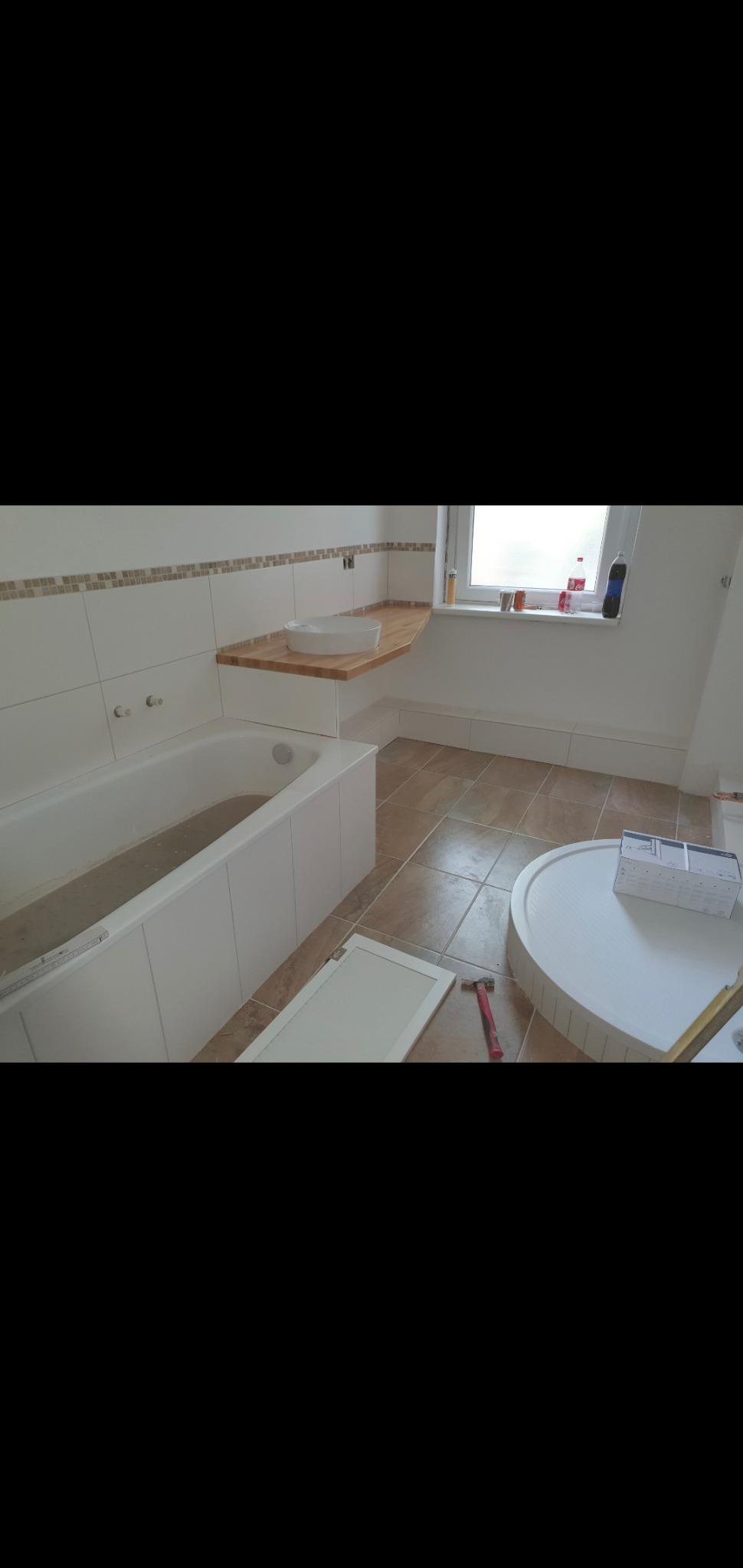Łazienka w trakcie remontu: biała wanna, drewniany blat pod umywalką, brązowe płytki podłogowe, niedokończona zabudowa wanny, narzędzia i materiały budowlane widoczne na podłodze.