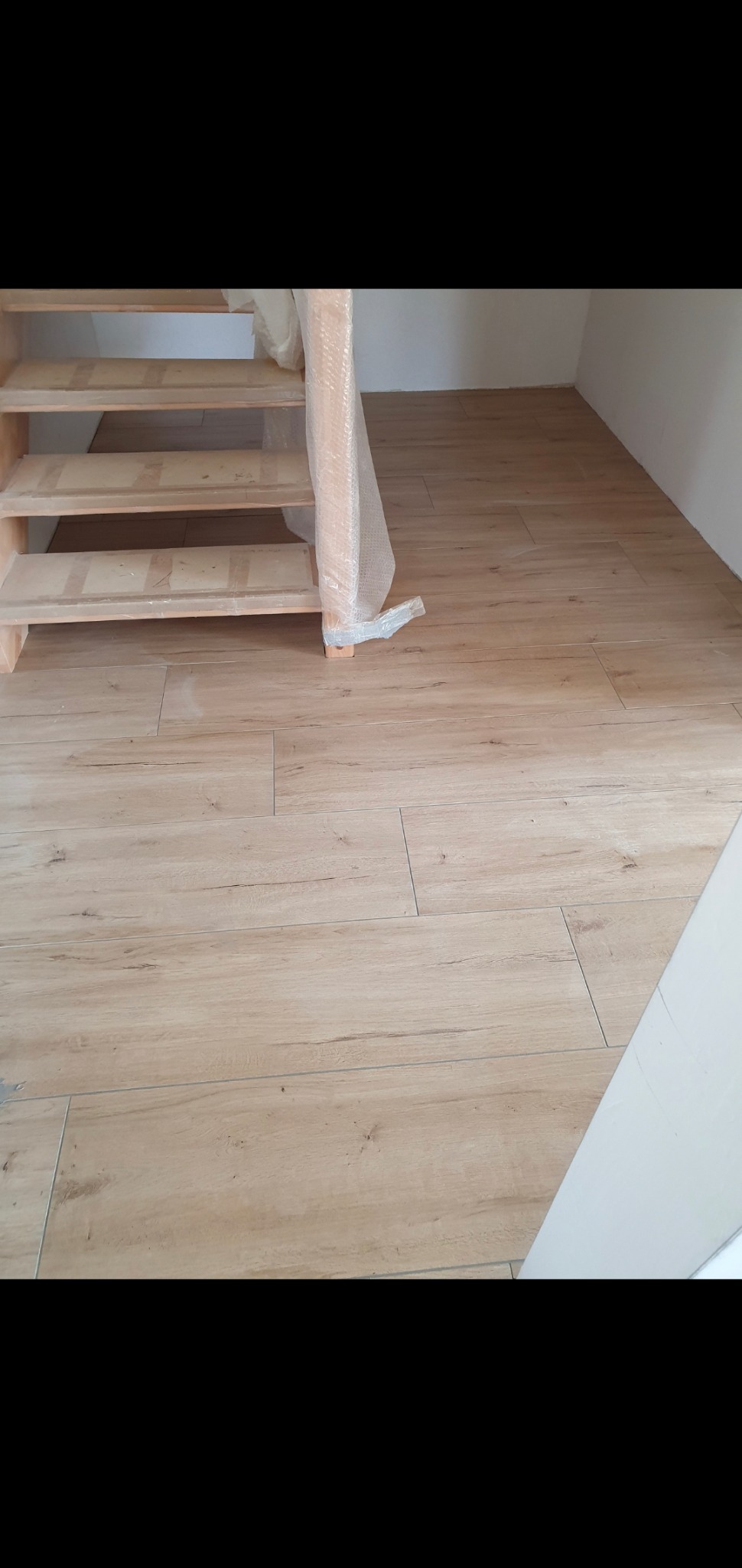 Jasne, drewnopodobne płytki podłogowe ułożone w pomieszczeniu, częściowo zakryte przez niezabezpieczone drewniane schody z folią ochronną.