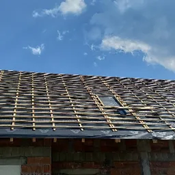 Konstrukcja dachu z łatami i kontrłatami pokryta membraną dachową, widoczne pojedyncze dachówki, budynek w trakcie budowy z czerwonej cegły, błękitne niebo z chmurami w tle.