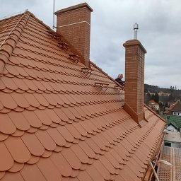 FIX-Dach - Montaż nowej dachówki ceramicznej w kolorze ceglastym, widoczne przygotowane podłoże z izolacją, komin z cegły i pracownik na dachu w tle, panorama miasta.