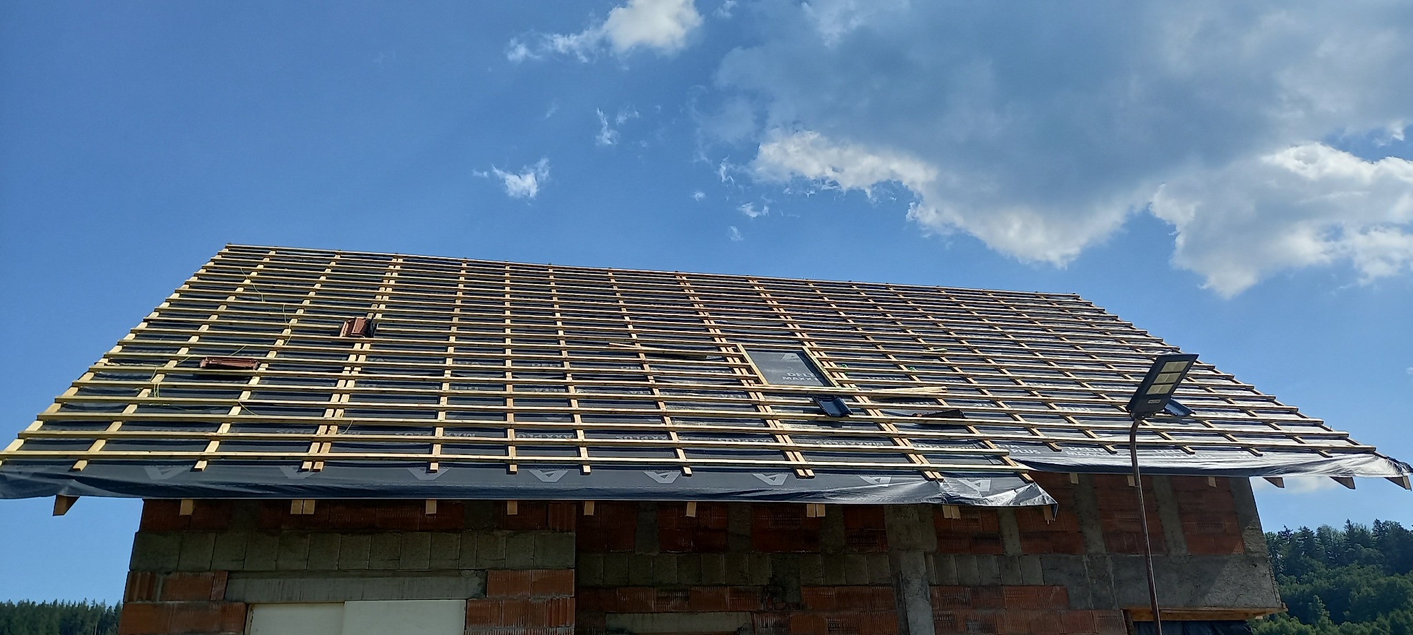 Konstrukcja dachu z łatami i kontrłatami pokryta membraną dachową, widoczne pojedyncze dachówki, budynek w trakcie budowy z czerwonej cegły, błękitne niebo z chmurami w tle.