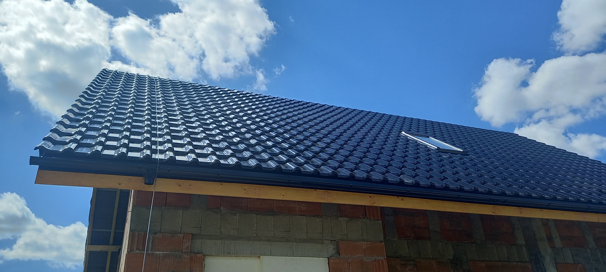 Nowy, błyszczący, grafitowy dach z oknem połaciowym, widoczny fragment muru z pustaków i niezakończone obróbki przy rynnie, na tle błękitnego nieba z białymi chmurami.