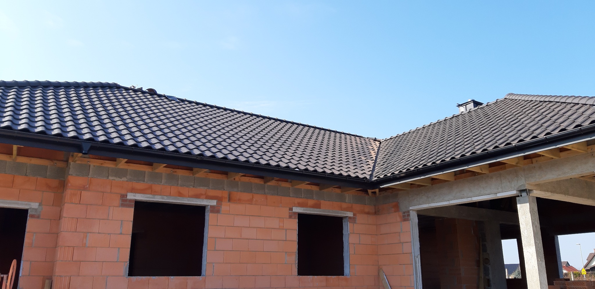 Nowy dach z ciemnej dachówki ceramicznej na budynku w stanie surowym otwartym, widoczne obróbki blacharskie i system rynnowy.