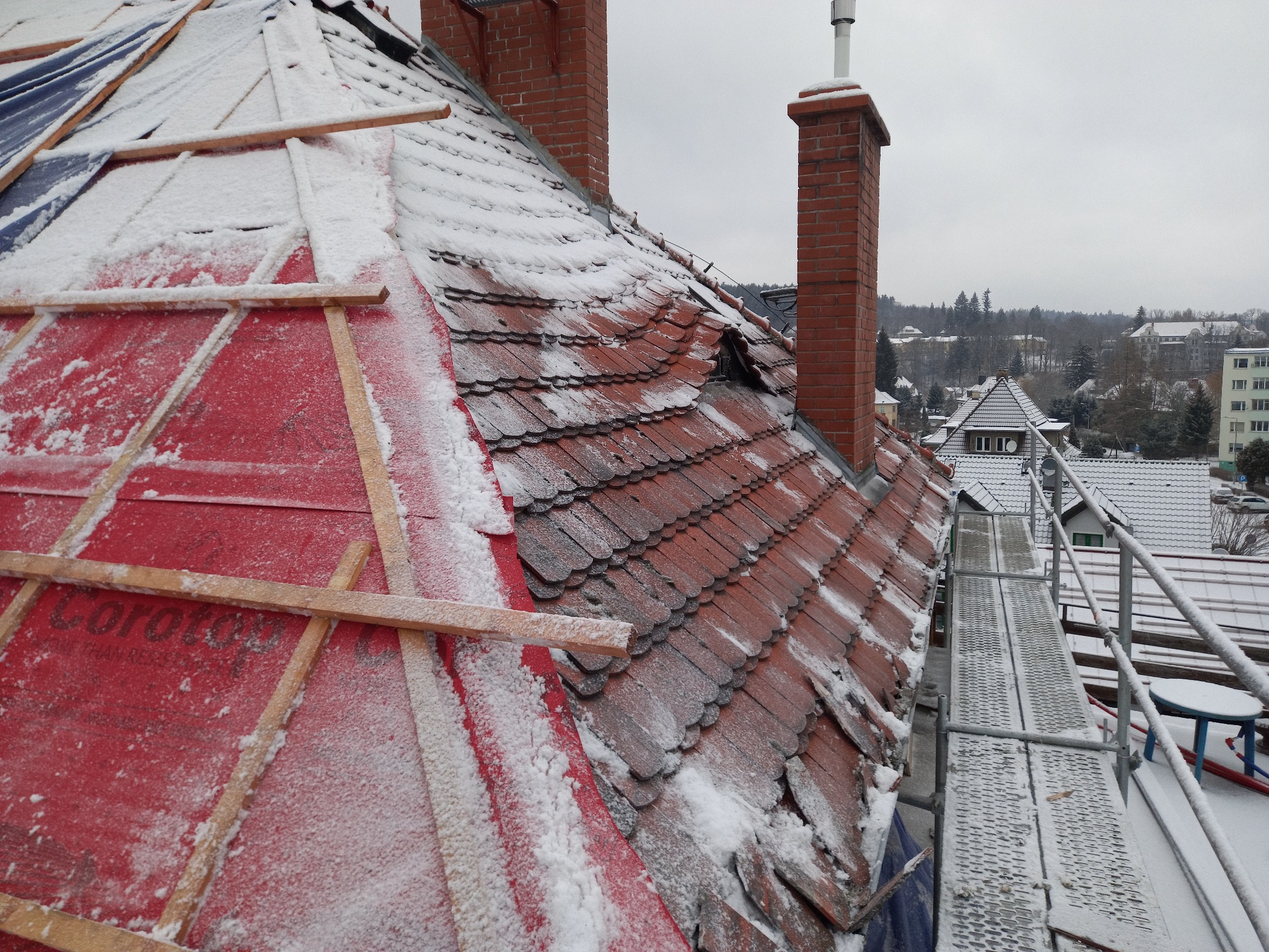 Śnieżny dach w trakcie remontu, widoczne czerwone membrany, fragmenty starej dachówki, kominy z cegły oraz rusztowanie