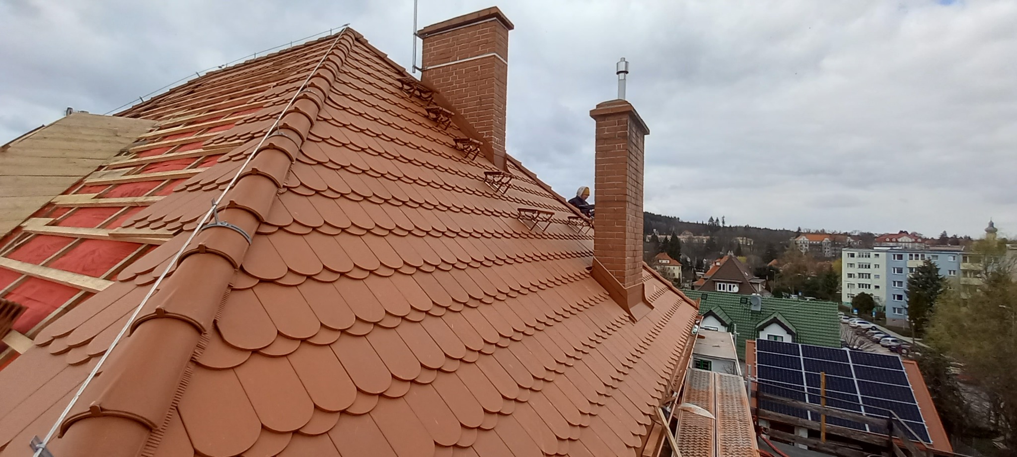 Montaż nowej dachówki ceramicznej w kolorze ceglastym, widoczne przygotowane podłoże z izolacją, komin z cegły i pracownik na dachu w tle, panorama miasta.