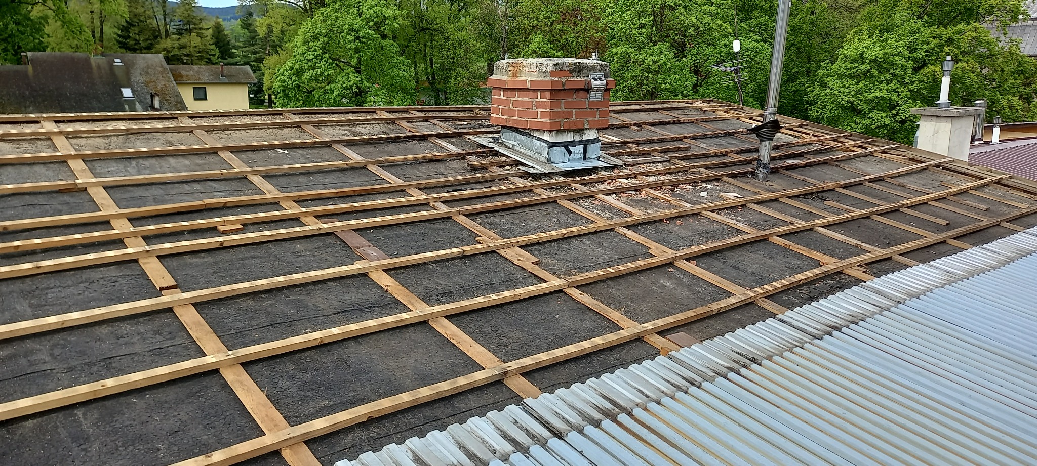 Dach w trakcie prac: widoczne łaty drewniane na czarnej papie, komin z cegły, elementy instalacji wentylacyjnej, fragment dachu z blachy falistej w tle drzewa.