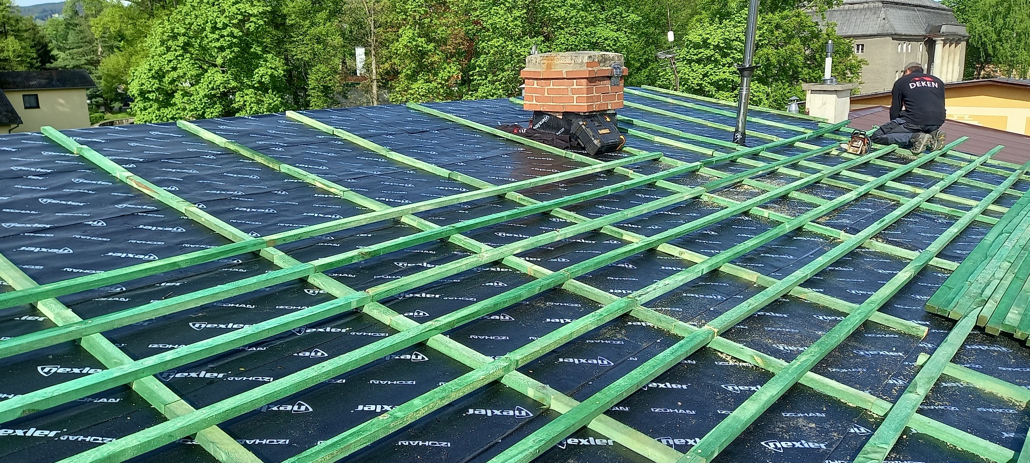 Konstrukcja dachu z zielonymi łatami i czarną membraną izolacyjną z widocznym kominem z cegły i pracownikiem w czarnym ubraniu na dachu budynku w tle.