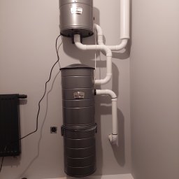 MONTER Andrzej Kosiewicz - Zamontowany, dwuczęściowy system centralnego odkurzania VacuMaid z rurami PCV na szarej ścianie, widoczne gniazdko elektryczne i fragment grzejnika.