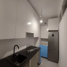 Wnętrze kuchni w trakcie montażu: białe szafki wiszące, czarny blat z poziomnicą, zlew z czarną armaturą, lodówka i niebieska szafka w tle.