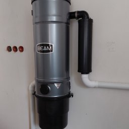 MONTER Andrzej Kosiewicz - Szary odkurzacz centralny marki BEAM zamontowany na białej ścianie, widoczne rury instalacyjne i niedokończone otwory elektryczne w ścianie.