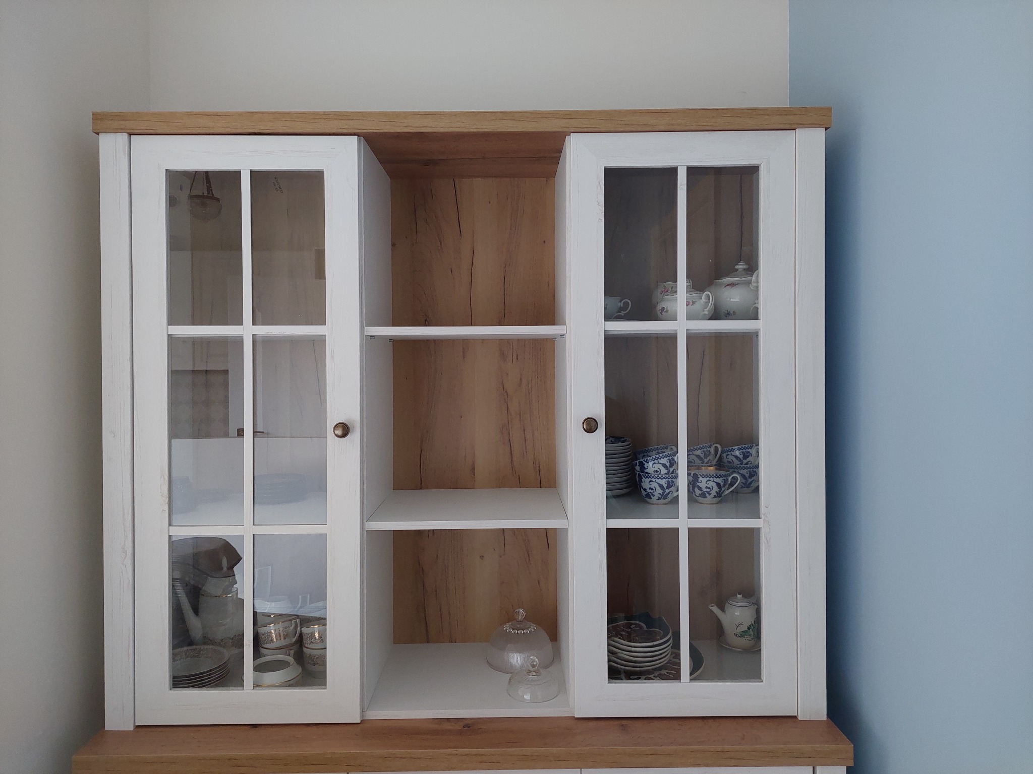 Biały kredens z przeszklonymi drzwiami, drewnianym blatem i wnętrzem, wypełniony porcelaną w różnych odcieniach błękitu i bieli, ustawiony na tle ściany w odcieniach bieli i błękitu.