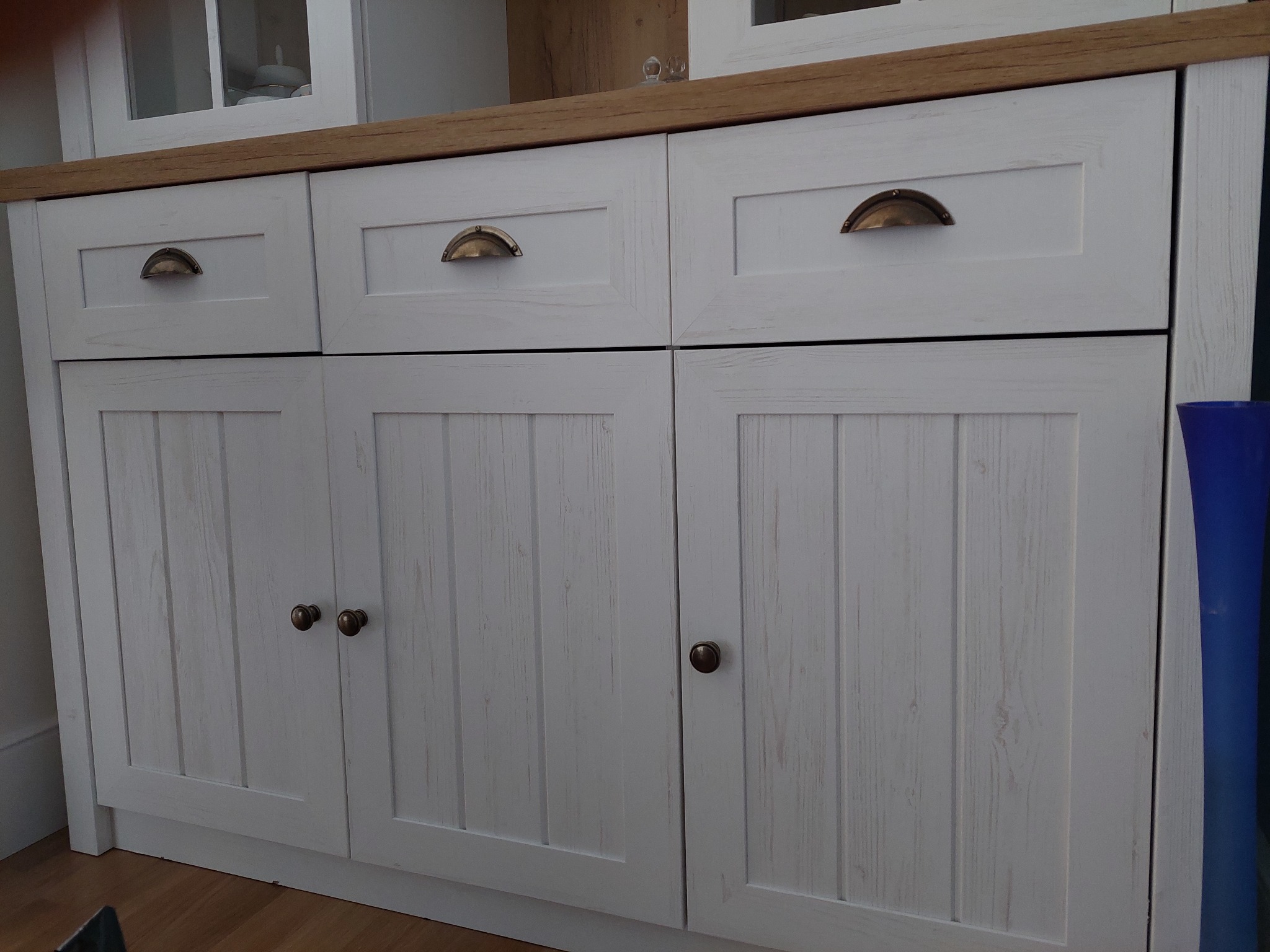 Biała drewniana komoda z trzema szufladami i trzema szafkami, z brązowymi uchwytami, stojąca na drewnianej podłodze, z widocznym fragmentem niebieskiego wazonu po prawej stronie.