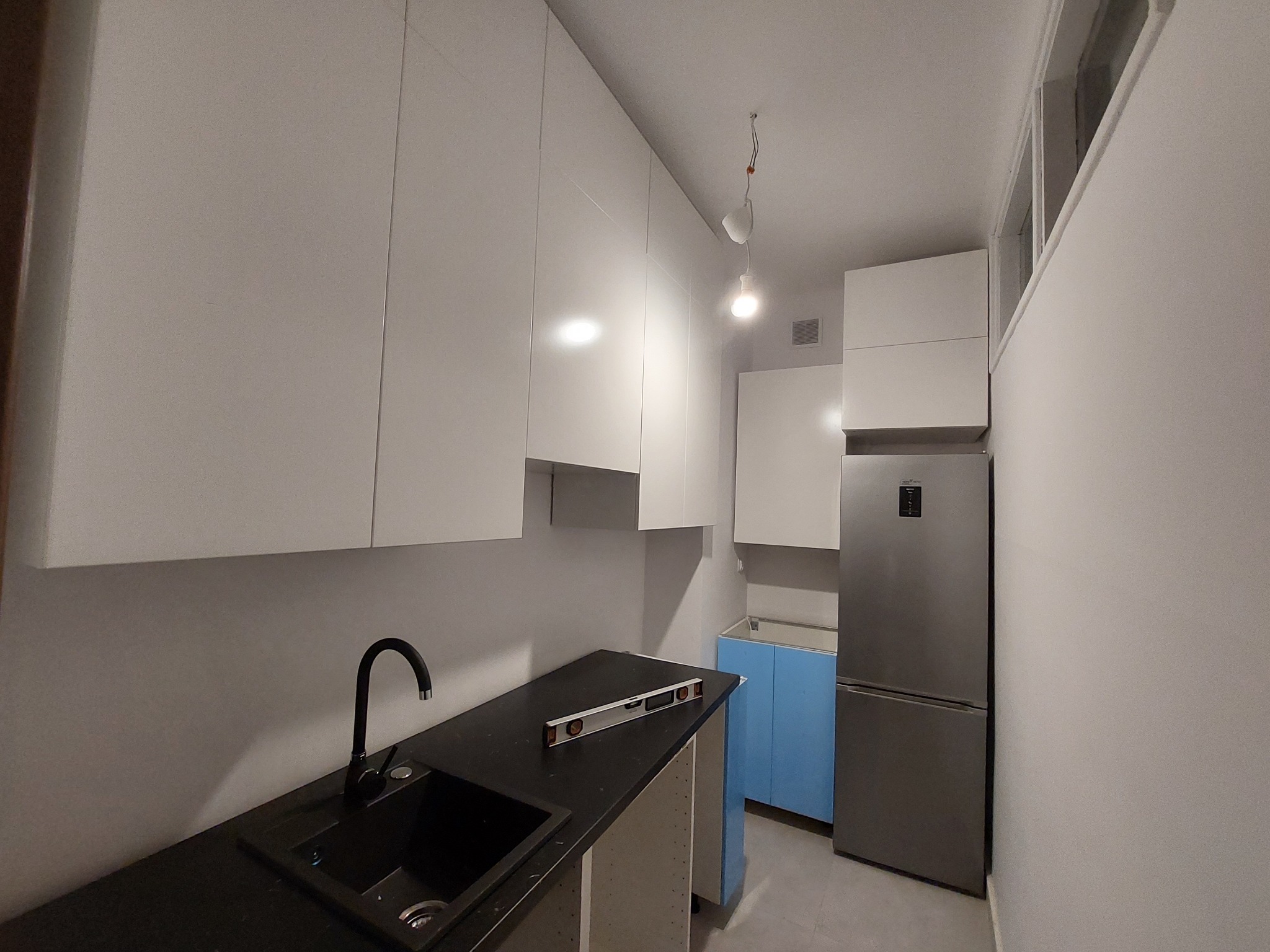 Wnętrze kuchni w trakcie montażu: białe szafki wiszące, czarny blat z poziomnicą, zlew z czarną armaturą, lodówka i niebieska szafka w tle.